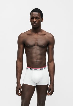 Modelo masculino de piel oscura y cuerpo tonificado vistiendo calzoncillos tipo bóxer blancos de Moschino con cintura negra y roja con marca, posando delante de un fondo liso y claro.