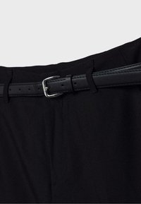 Ceinture noire avec boucle argentée passant dans les passants d’un pantalon noir, centrée sur un fond clair uni.