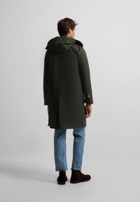 Parka verde oscuro con capucha, cremalleras y puños abotonados. El diseño tiene un corte largo y una parte trasera dividida, combinado con jeans azul claro y botas marrones.