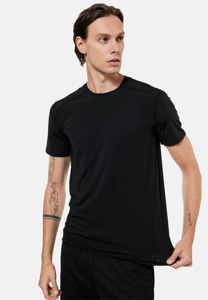 Mies pukeutunut mustaan urheilupaitaan ja shortseihin, vetää hieman paidan helmaa, molemmissa käsivarsissa tatuoinnit, katsoo vasemmalle.