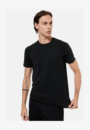 Man wearing black athletic t-shirt and shorts, pulling shirt hem slightly, with tattoos on both forearms, looking to his left.