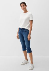 Frau trägt ein weißes Kurzarmhemd, eine blaue verkürzte Jeans mit seitlichen Schlitzen und weiße Turnschuhe, steht vor einem schlichten hellen Hintergrund.