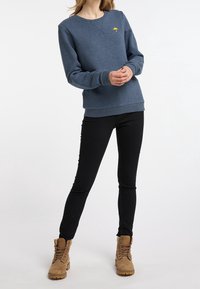 Blaues Sweatshirt mit gesticktem gelbem Regenschirm-Logo, kombiniert mit schwarzen Skinny Jeans und beigen Schnürstiefeln. Lässiger, bequemer Schnitt.