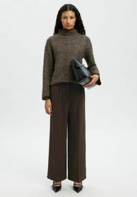 Femme portant un pull marron à motif chevron, un pantalon large marron, des chaussures pointues noires, tenant un sac à main en cuir noir sur un fond blanc.