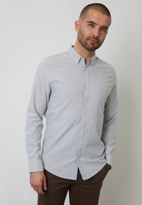 Camisa abotonada gris claro en una tela suave, con mangas largas, cuello clásico y un sutil acento de logo. Llevada con pantalones marrones.