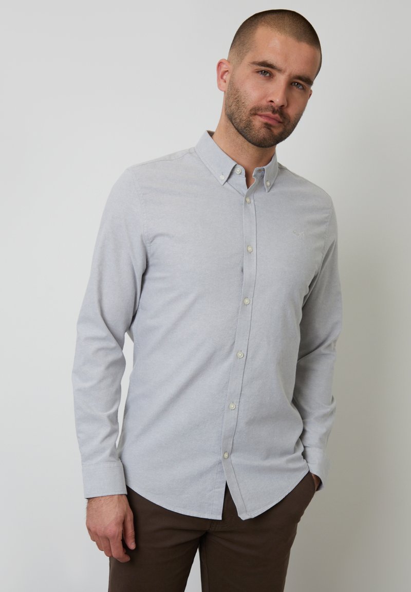 Camisa abotonada gris claro en una tela suave, con mangas largas, cuello clásico y un sutil acento de logo. Llevada con pantalones marrones.