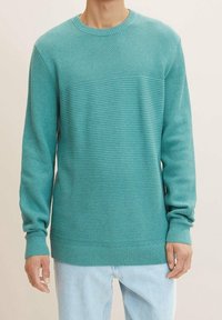 Homme portant un pull en maille texturée turquoise avec des poignets et un ourlet côtelés, associé à un jean bleu clair, sur un fond uni.