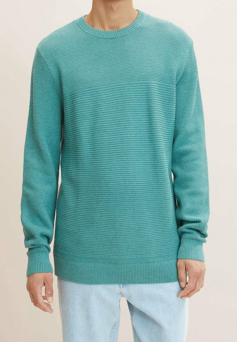 Homme portant un pull en maille texturée turquoise avec des poignets et un ourlet côtelés, associé à un jean bleu clair, sur un fond uni.