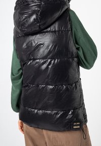 Schwarze Steppweste mit glänzendem, gesteppte Außenseite und verstellbarer Kapuze, getragen über einem grünen Kleidungsstück. Verfügt über eine Seitentasche und Markenschild am Saum.