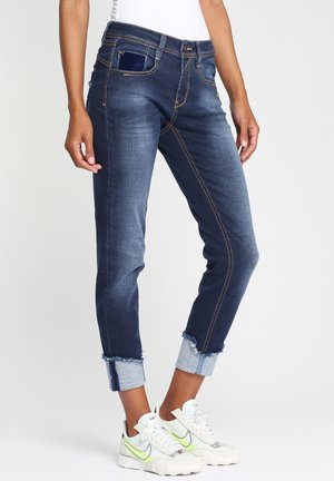 Persoon draagt donkerblauwe denim jeans met omgeslagen pijpen en witte sneakers, staat met een telefoon zichtbaar in de voorzak.