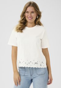Weißes Baumwoll-T-Shirt mit kurzen Ärmeln, gewelltem Saum und floraler Stickerei am Saum. Kombiniert mit hellblauen Jeans.