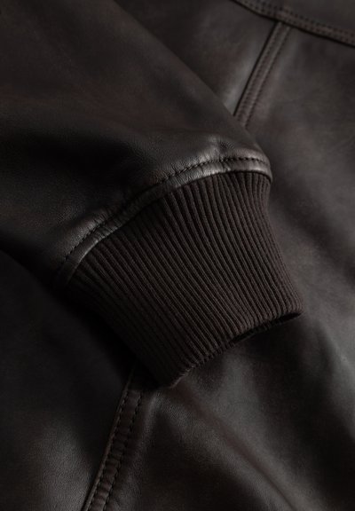Közelkép egy sötétbarna bőrkabát ujjáról, bordázott kötött mandzsettával és látható varrási részletekkel.