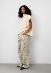 Sukienka bez rękawów w kolorze kremowym z wysokim kołnierzem, zestawiona z luźnymi spodniami w marmurkowy wzór w odcieniu beżowym i czarnymi, spiczastymi balerinami.