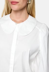 White collared shirt with a button at the neck, featuring puffed sleeves and smooth fabric texture. Silver earrings visible in the foreground.