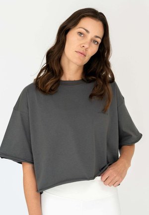 Frau mit schulterlangem braunem Haar, die ein übergroßes dunkelgraues T-Shirt und weiße Hose trägt, steht vor einem einfarbigen Hintergrund.