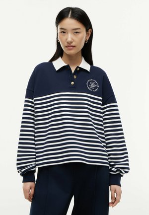 Jonge vrouw die een marineblauw rugbyshirt draagt met witte horizontale strepen, een witte kraag en gouden knopen, staand tegen een effen achtergrond.