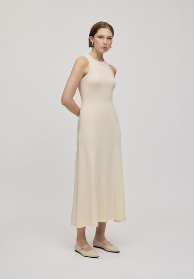 Abito maxi beige senza maniche realizzato in tessuto morbido, aderente nella parte superiore e svasato verso il fondo, abbinato a scarpe chiare.