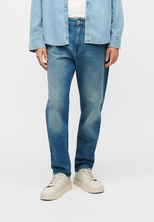 Personne portant un jean en denim bleu clair, une chemise en denim claire par-dessus un haut blanc, et des baskets blanches, debout devant un fond uni.