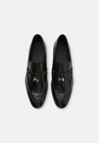 Mocasines negros de cuero con grabado de cocodrilo, con detalles de borlas y herrajes en tono dorado. Textura suave y diseño de perfil bajo.