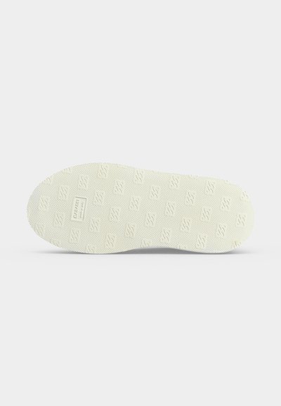 Semelle d'une chaussure en caoutchouc blanche avec un motif texturé de petits logos en relief et une étiquette de marque rectangulaire près du talon.