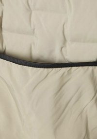 Beige gepolstertes Gewebe mit einer glatten Textur, das an der Oberseite eine schwarze Nahtkante aufweist. Die Oberfläche zeigt leichte Falten und Knicke.