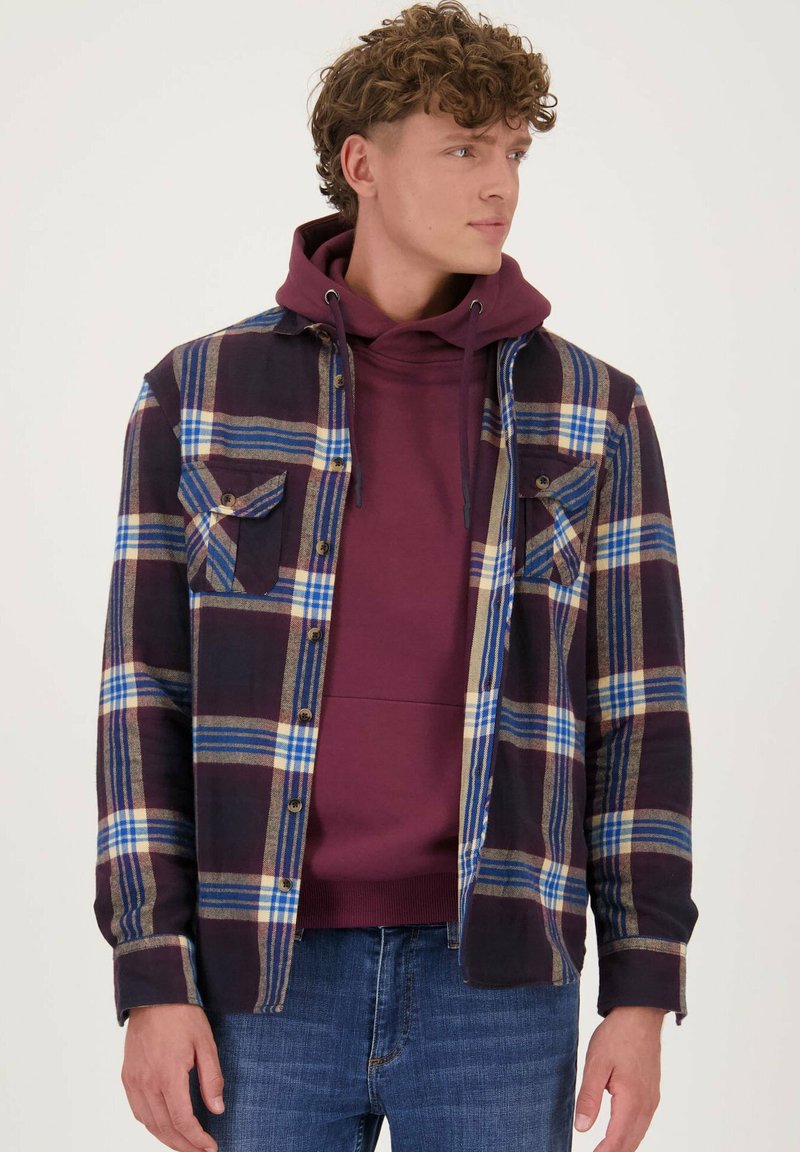 Geruit flanellen shirt in donkerpaars, blauw en beige, gelaagd over een bordeauxrode hoodie. Beschikt over twee borstzakken en knoopsluiting.