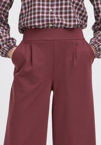 Pantaloni largi bordo cu talie înaltă, prezentând buzunare laterale și detalii cu pliu pentru un fit structurat. Material neted, elastic.
