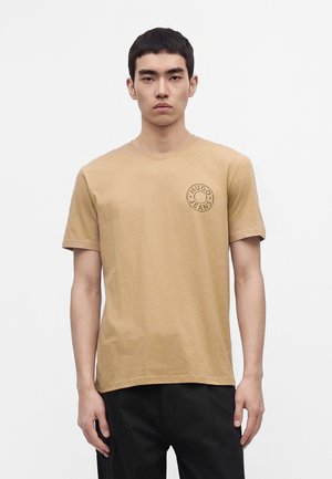 Jeune homme aux cheveux noirs courts portant une chemise à manches courtes beige Hugo Jeans et un pantalon noir, debout devant un fond blanc uni.