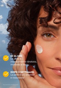 Primo piano di una mano che applica delicatamente una crema bianca su una guancia. Sullo sfondo si vede un cielo blu con nuvole. La pelle appare liscia e idratata.