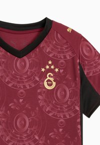 Burgundin urheiluverkko V-kaula-aukolla, mustilla yksityiskohdilla, kultaisella logolla, viidellä tähdellä ja teksturoiduilla pyöreillä kuvioilla kankaassa.