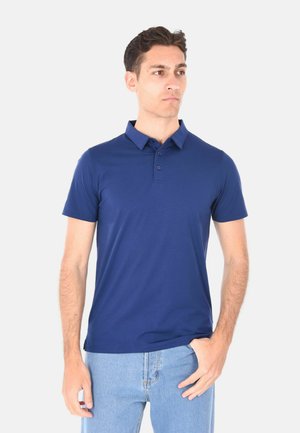 Giovane uomo che indossa una polo aderente blu scuro a maniche corte e jeans azzurri, in piedi con una mano in tasca, che guarda di lato.