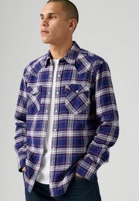 Camicia a maniche lunghe a scacchi blu e bianchi con taschini sul petto e chiusura con bottoni. Il tessuto sembra morbido; presenta linee rosse a contrasto.