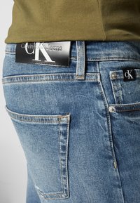 Ljusblå denimjeans med en urblekt textur, med en svart Calvin Klein-logotyp på midjebandet och en liten logotags på fickan.