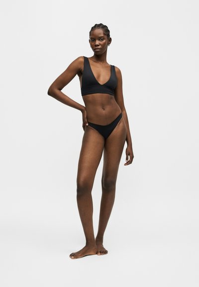 Γυναίκα στέκεται ξυπόλυτη με το χέρι στη μέση, φορώντας μαύρο μπικίνι μπροστά σε απλό γκρι φόντο.