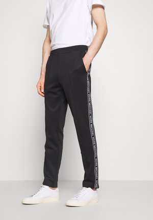 Muž nosící černé tepláky s bílými proužky s logem "Michael Kors" po stranách, bílé tenisky a bílé krátké tričko.