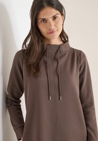 Sweat à capuche marron avec un col montant et des cordons, présentant une texture lisse et des manches longues. Comprend de petits accents en métal au niveau du col.