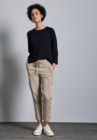 Marine gebreide trui met een losse pasvorm, gecombineerd met beige cargobroeken met zijzakken. Model draagt witte sneakers; neutrale achtergrond.
