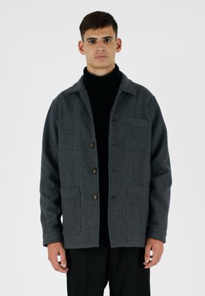 Veste en laine gris foncé avec un col, fermeture à boutons, deux poches avant et une coupe décontractée. Portée sur un col roulé noir.