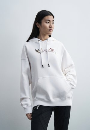Personne portant un sweat à capuche blanc avec des motifs d'oiseaux et le texte « Off-White », debout devant un fond gris clair uni.