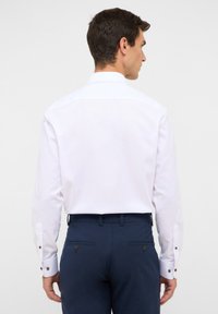Camicia bianca a maniche lunghe con colletto standard, dotata di polsini con bottoni, abbinata a pantaloni blu scuro con tasche posteriori e chiusure a bottone.