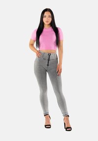 Nainen, jolla on pitkä musta tukka ja joka on pukeutunut vaaleanpunaiseen lyhyeen t-paitaan, korkeavyötäröisiin harmaisiin kapeisiin farkkuihin ja mustiin korkeakorkoisiin sandaaleihin, seisoo käsi lanteilla.