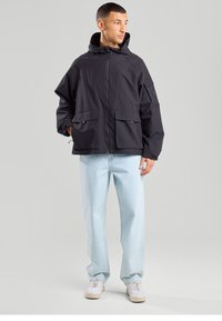 Black waterproof jacket with a hood, front zip, two large front pockets, and loose fit, paired with light blue jeans and white sneakers.