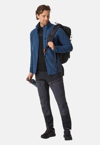 Niebieska polarowa kurtka, czarny t-shirt, ciemne spodnie cargo z zamkami, czarny plecak z pomarańczowym zapięciem i czarne sneakersy; trzyma butelkę wody.