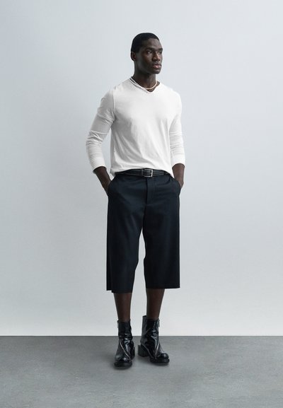 Άνδρας που φορά λευκό πουκάμισο με μακριά μανίκια, μαύρο φαρδύ παντελόνι μέχρι τον αστράγαλο, μαύρες μπότες με τακούνι και κολιέ με χάντρες, στέκεται μπροστά σε απλό γκρι τοίχο.