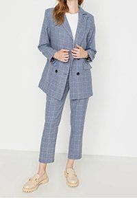 Blazer à carreaux bleu avec deux boutons sur le devant, associé à un pantalon assorti et des mocassins beige. Tissu texturé et design structuré sont remarquables.