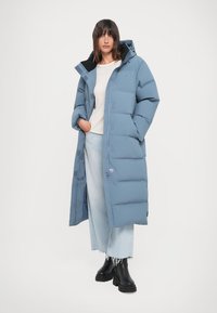 Light blue long puffer coat with a zip front, a hood, and side pockets; paired with light jeans and black platform boots.