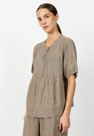 Frau mit dunklem, zurückgebundenem Haar, die ein lockeres, kurzärmeliges beiges Leinenoberteil und passende Hose trägt, vor einem schlichten weißen Hintergrund stehend.