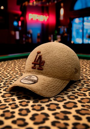 Beige textured cap with brown "LA" logo and adjustable sticker, placed on a leopard print surface with blurred colorful background lights.