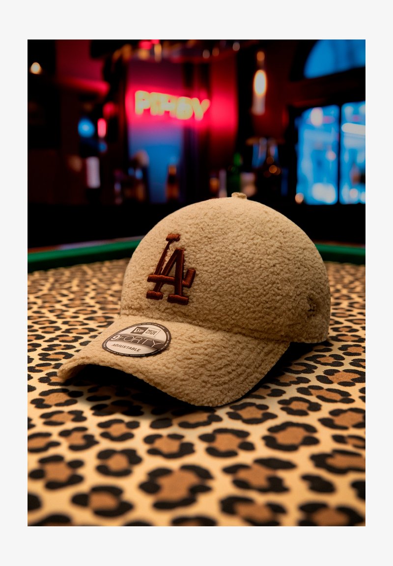 Beige teksturēta cepure ar brūnu "LA" logotipu un regulējamu uzlīmi, novietota uz leoparda raksta virsmas ar izplūdušiem krāsainiem fona gaismas efektiem.