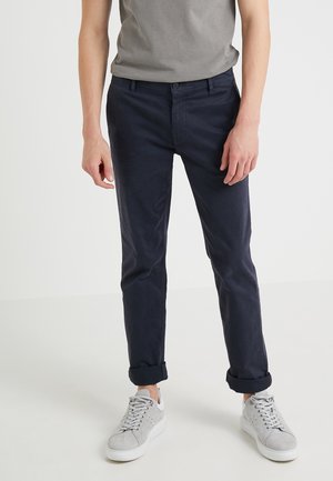 Person trägt dunkelblaue Chino-Hose, an den Knöcheln hochgekrempelt, und hellgraue Sneaker vor einem einfachen weißen Hintergrund.
